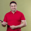 delivery man wearing red uniform and cap holding clipboard looking positive and happy smiling standing over isolated green background